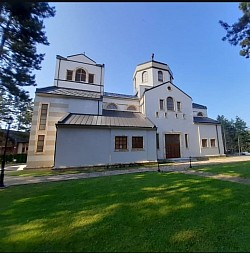The magnificent Temple of the Transfiguration on Zlatibor, shot from a distance, shows monumental architecture in the Raška style with white facade walls and distinctive red roofs, surrounded by a landscaped promenade and tall pine trees under the clear blue sky of the mountain tourist center