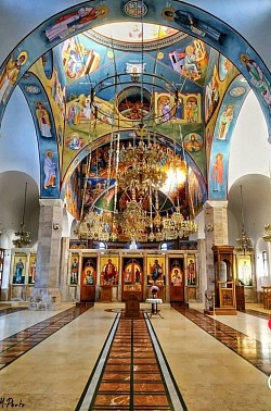 The magnificent Temple of the Transfiguration on Zlatibor, shot from a distance, shows monumental architecture in the Raška style with white facade walls and distinctive red roofs, surrounded by a landscaped promenade and tall pine trees under the clear blue sky of the mountain tourist center
