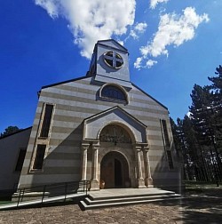 View of the Church of the Transfiguration of the Lord from a landscaped path in the park where the entire northern facade with the bell tower and central dome, which dominate the surrounding mountain landscape, can be seen through the treetops.